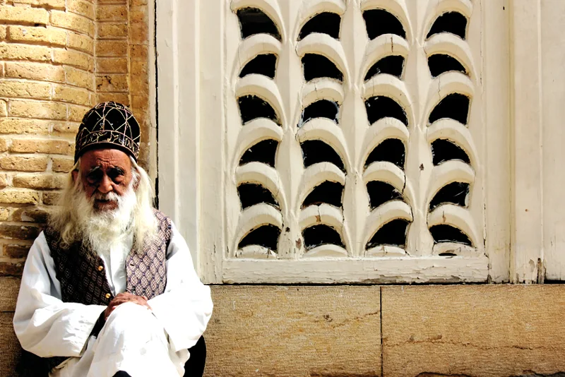 An older man seated beside an ornate brick and plaster wall